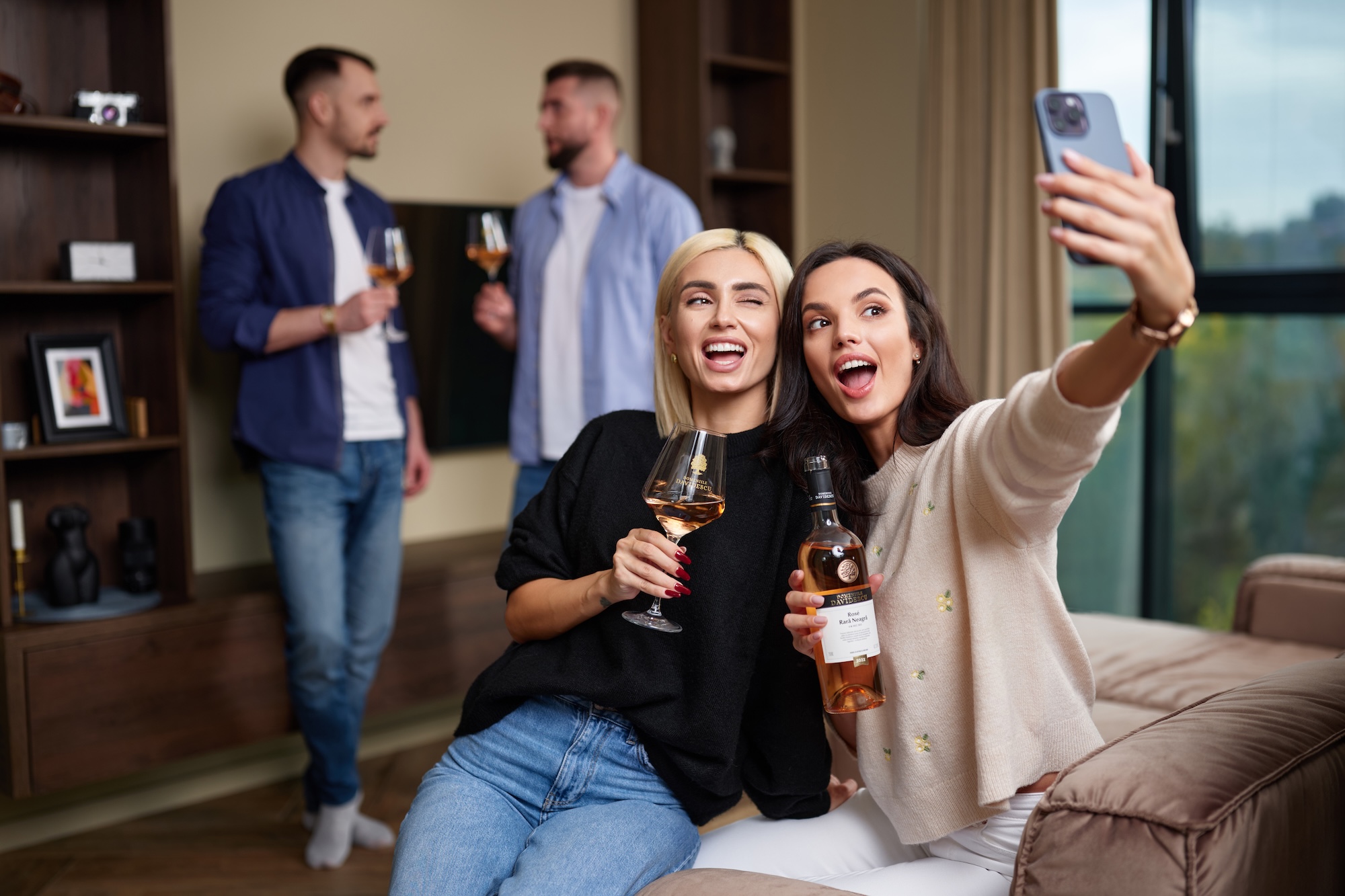 Doua femei isi fac selfie cu vin rose, intr-o sufragerie