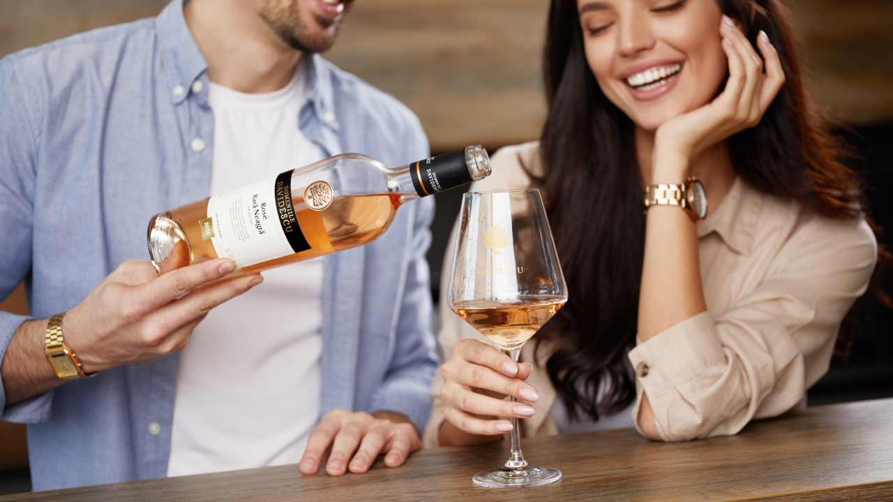 Cuplu degustând vin rose într-un bar, bărbatul toarnă vin într-un pahar, iar femeia zâmbește